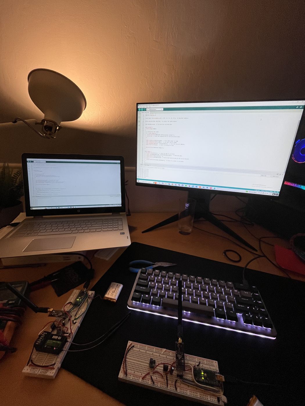 The development bench: both halves of the relay wired on breadboards, firmware open in the Arduino IDE on two screens.
